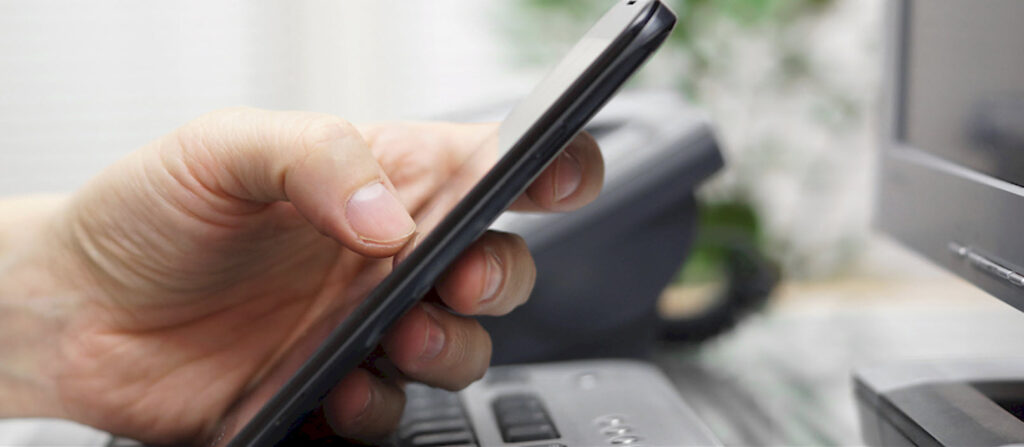 Hand holding smartphone near a computer, representing communication with WordPress experts for inquiries and support at Secure WP Solutions.